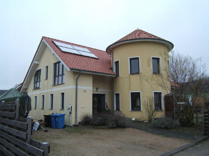 Zweifamilienhaus in Hildesheim, Langer Kamp 26, mit gelber Fassade, rotem Satteldach, runder Erweiterung und Solaranlage auf dem Dach. Vorgarten mit Pflanzen und Mülltonnen