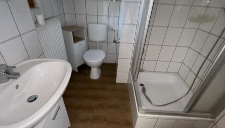 Badezimmer mit weiß gefliesten Wänden, WC, Dusche mit Glastür, Waschbecken und Holzboden. Standort: Burgstraße 11, 07607 Eisenberg