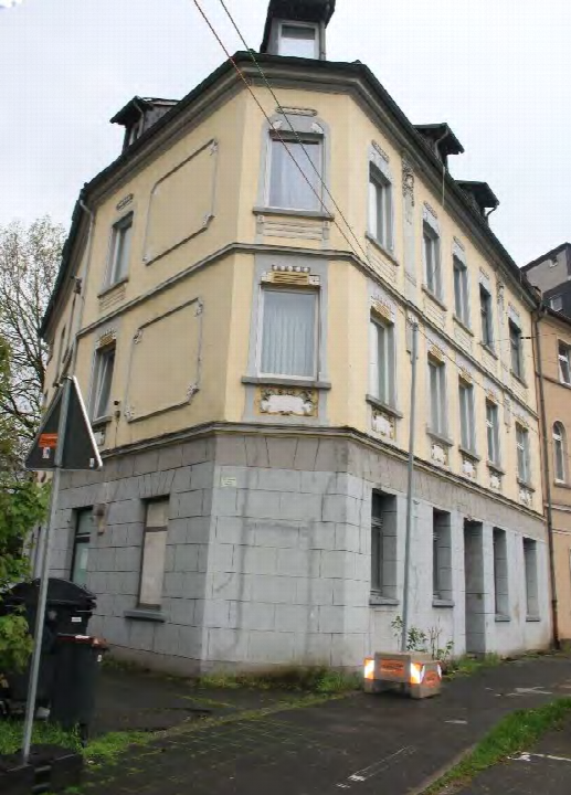 Eckansicht eines Mehrfamilienhauses mit gelber Fassade, grauem Sockel und Dachgauben. Die Fassade zeigt Fenster mit Verzierungen und eine Straßenecke in Castrop-Rauxel