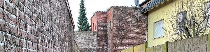 Seitenansicht eines Backsteinhauses mit angrenzenden Mauern, einem Holzzaun und kahlen Bäumen im Vordergrund. Im Hintergrund ist ein Nadelbaum sichtbar