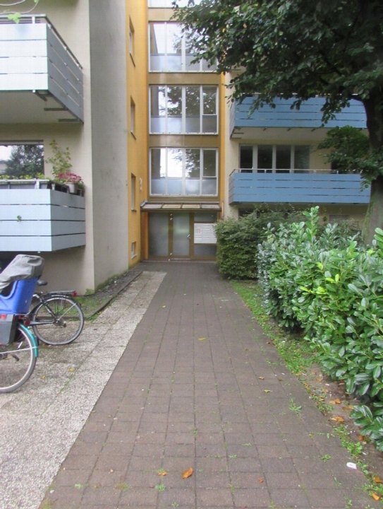 Eingangsbereich eines Mehrfamilienhauses mit gepflastertem Weg, Balkonen und Grünanlagen. Gebäude in Gelb- und Blautönen