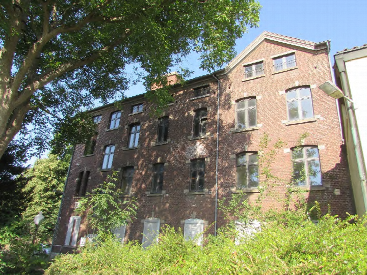 Seitenansicht eines mehrstöckigen Backsteinhauses mit Fenstern und umgebenden Bäumen und Sträuchern in Bergheim, Pützer Straße 9