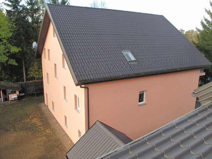 Seitenansicht eines mehrstöckigen Gebäudes mit rosa Fassade und dunklem Wellendach, umgeben von Bäumen und einem gepflasterten Hof