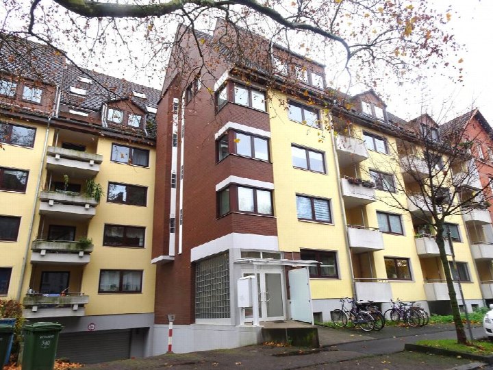 Eingangsbereich eines Mehrfamilienhauses mit weißer Tür, Glasblockwand und Vordach, umgeben von gelb- und braun verputzten Fassadenabschnitten