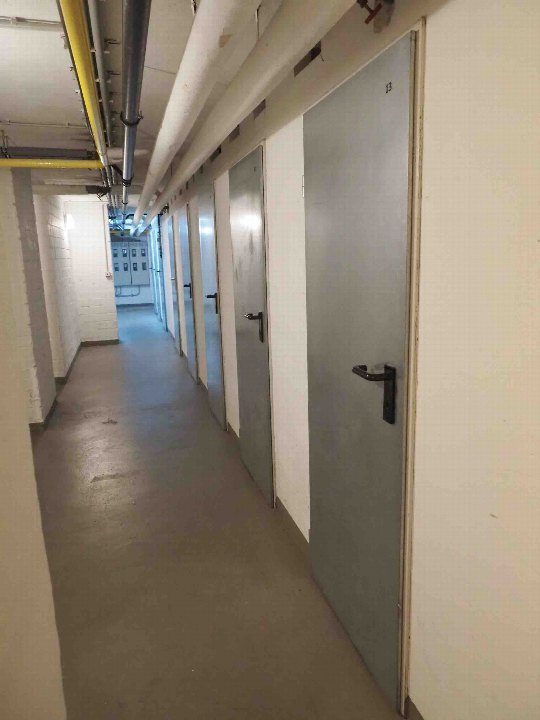 Kellerflur mit Betonboden, Metalltüren und sichtbaren Rohrleitungen an der Decke