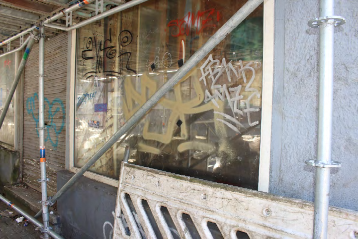 Seitenansicht eines Gebäudes mit Graffiti an der Fensterscheibe, Baugerüst und Holzbarriere im Vordergrund