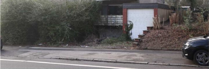 Garage mit weißer Tür und Ziegelrahmen, Zugang über Stufen, umgeben von Vegetation und Mulchhaufen, geparktes Fahrzeug rechts