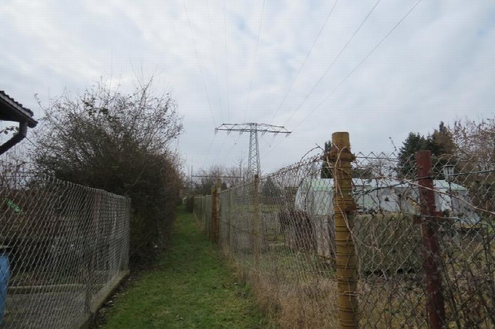 Grasbewachsener Weg zwischen Maschendrahtzäunen, Sträuchern und Strommasten im Außenbereich der Immobilie Altenburg 44