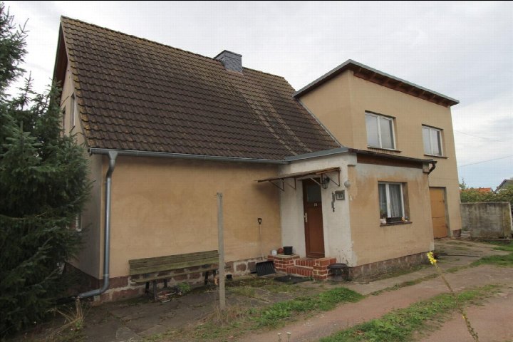 Einfamilienhaus mit braunem Ziegeldach und beigefarbener Fassade, Eingangsbereich mit Vordach und Gartenbereich vorne
