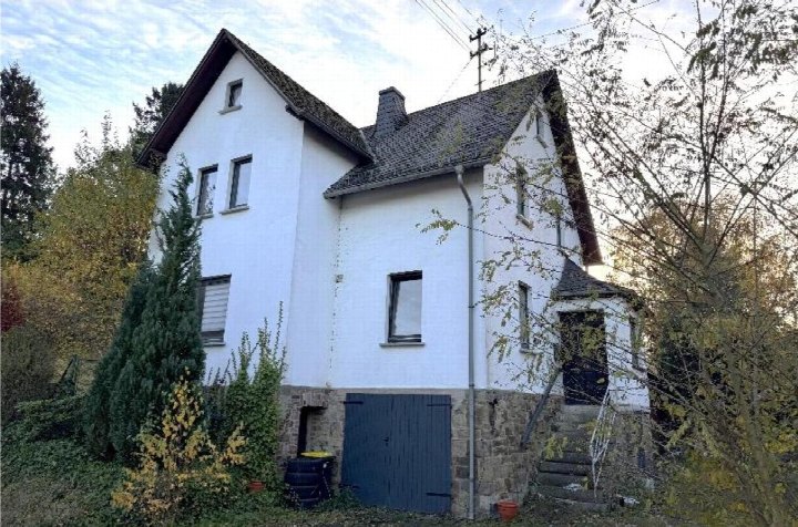 Seitenansicht eines zweistöckigen Einfamilienhauses mit weißer Fassade, Steinsockel, dunklem Dach und umgebenden Bäumen und Sträuchern