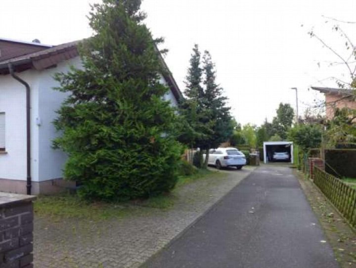 Auffahrt mit gepflastertem Weg zu einer Garage, daneben ein weißes Fahrzeug und ein großer Nadelbaum vor einem weißen Gebäude