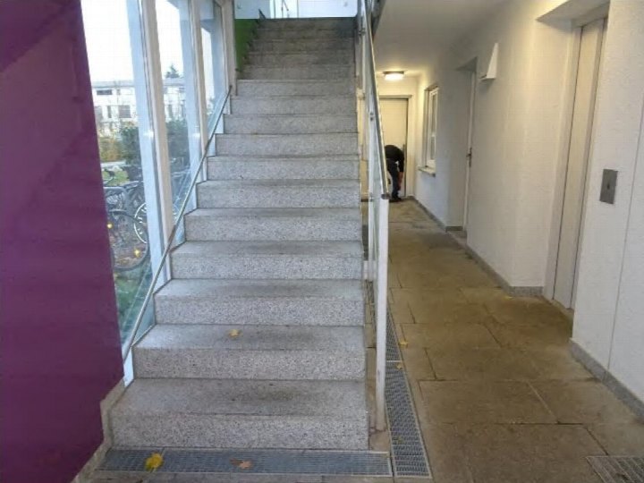 Treppenhaus mit Betontreppen und Geländern, rechts Flur mit Türen, links Fensterfront mit Blick nach draußen