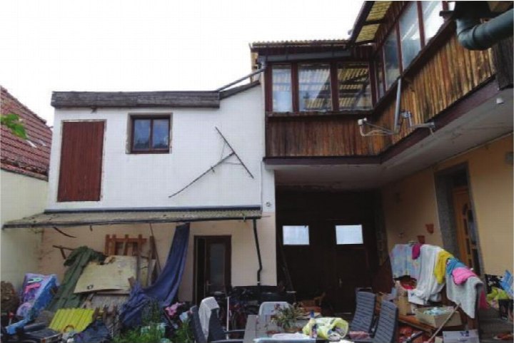 Rückansicht eines Einfamilienhauses mit Balkon, Fenstern und unordentlichem Hofbereich mit Gegenständen und Wäsche