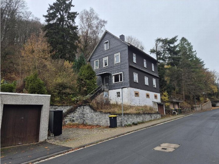 Seitenansicht eines zweistöckigen Hauses mit dunkler Fassade und weißem Sockel, umgeben von Bäumen und einem Steinmauerwerk, am Hang in Biedenkopf