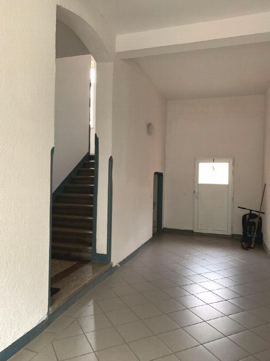 Treppenhaus mit weiß gestrichenen Wänden, Fliesenboden und Treppe nach oben, Eingangstür im Hintergrund