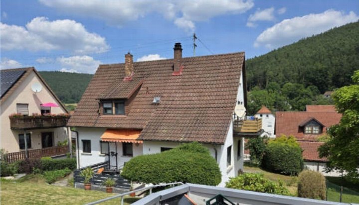 Einfamilienhaus mit Satteldach und weißer Fassade, Vorgarten, Eingang mit Markise, im Hintergrund bewaldeter Hügel