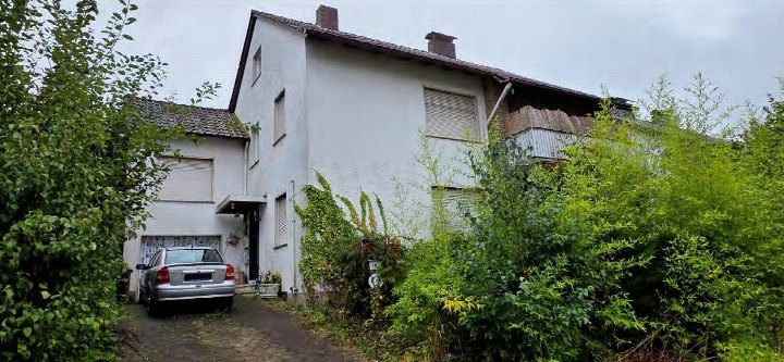Seitenansicht eines zweistöckigen Wohnhauses mit Ziegeldach, Fenstern und umgebenden Sträuchern. Ein silberner PKW steht auf dem Kiesweg vor der Garage