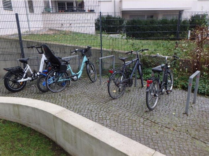 Vier Fahrräder stehen an einem Metallständer auf einem Kopfsteinpflaster vor einem Zaun und einem Gebäude in Berlin
