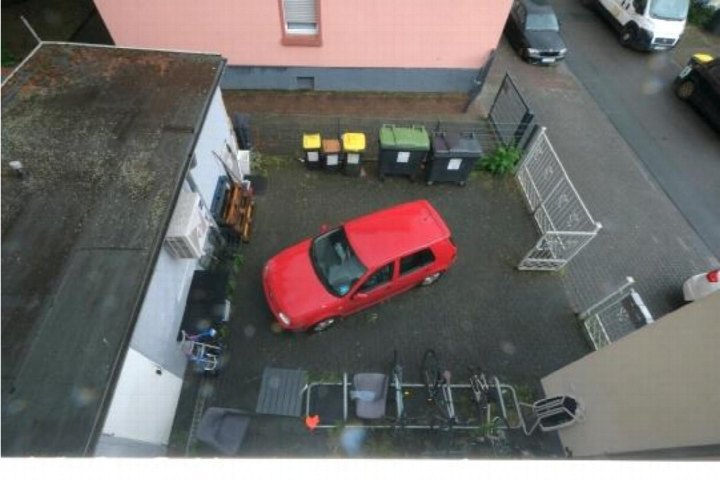 Überdachter Parkplatz mit rotem Auto, Müllcontainern und Metallzaun neben einem Wohn-/Geschäftshaus in Frankfurt am Main