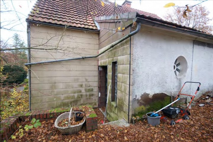 Seitenansicht eines Einfamilienhauses mit Ziegeldach, verputzten und verkleideten Wänden sowie Gartenbereich mit Laub und Gartengeräten