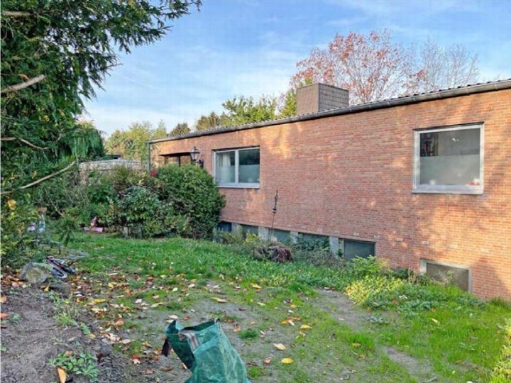 Gartenbereich mit Gras, Sträuchern und Laub neben einem Backsteinhaus, teilweise mit Gartengeräten und einem grünen Tarp