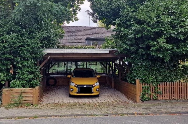 Gelber Wagen unter Holzcarport mit Gravelboden, umgeben von grüner Hecke und Holzzaun, im Vorgarten eines Einfamilienhauses