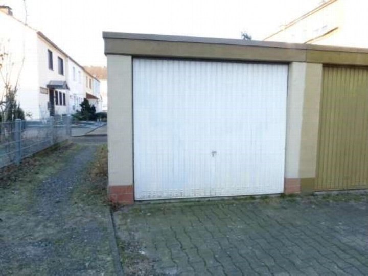 Garage mit weißer Rolltür und angrenzender Wand, gepflasterter Vorplatz, im Hintergrund Reihenhaus und Zaun