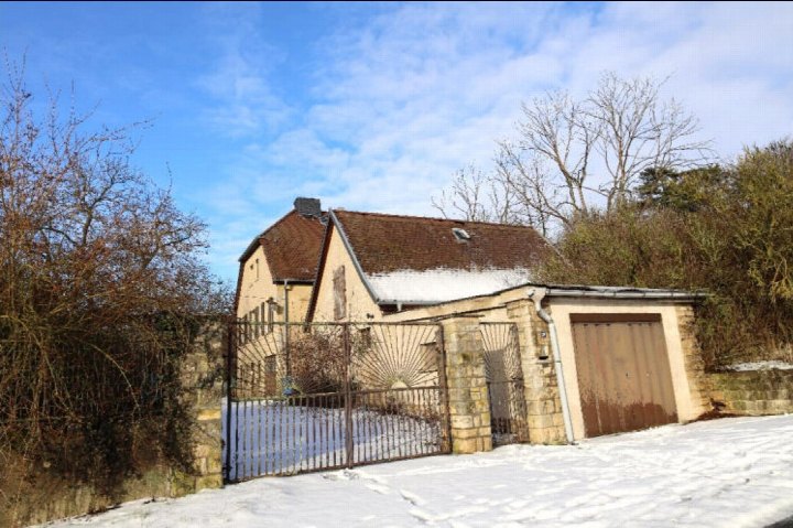 Einfamilienhaus mit Ziegeldach, Einfahrtstor und Garagenanbau, verschneiter Vorgarten, kahle Bäume im Hintergrund