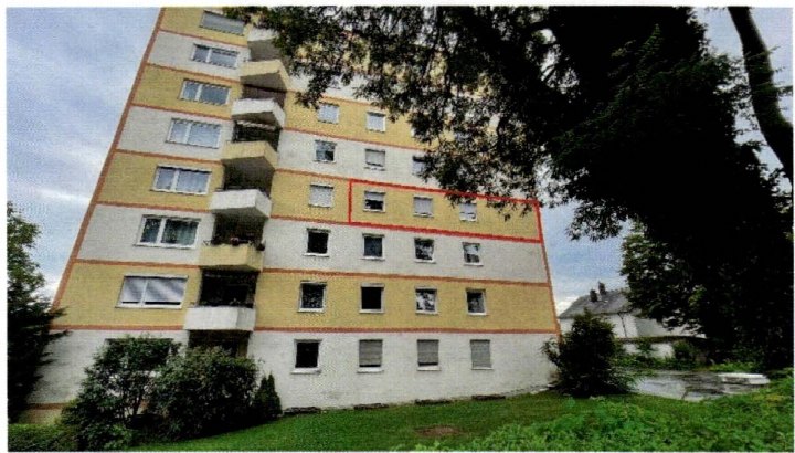 Seitenansicht eines Mehrfamilienhauses mit gelb-weißer Fassade, Balkonen und umgebenden Bäumen und Grünflächen