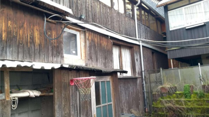 Rückansicht eines verwitterten Holzhauses mit Basketballkorb und eingezäuntem Gartenbereich in Schalkau