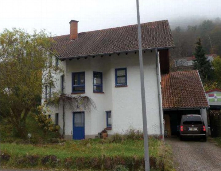 Einfamilienhaus mit weißer Fassade, blauen Fensterrahmen und Ziegeldach, Vorgarten und Garage mit geparktem Fahrzeug