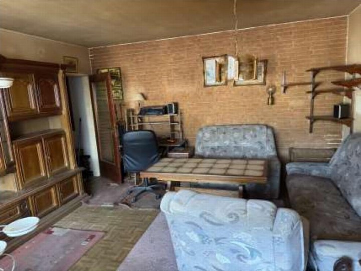 Wohnzimmer mit gemusterten Sofas, Holztisch, Schreibtischstuhl und Holzschrank, Backsteinwand im Hintergrund