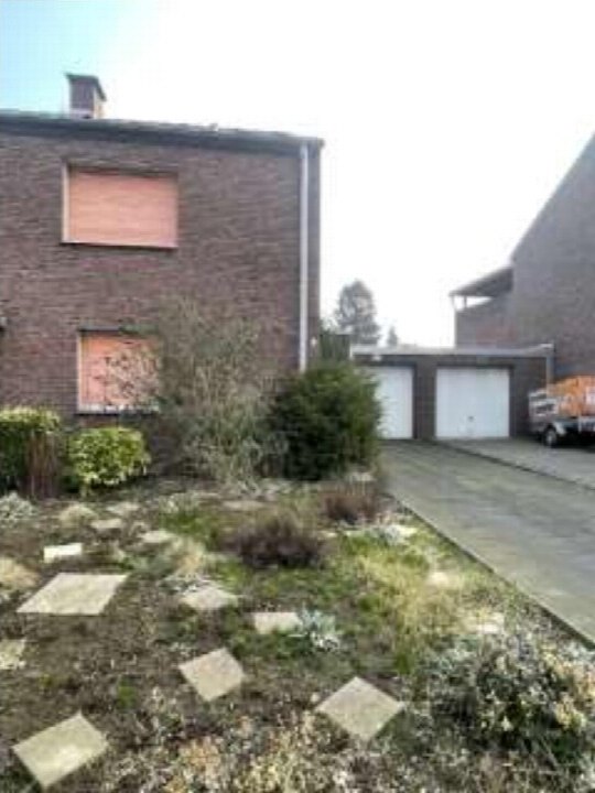 Überwuchertes Grundstück mit Steintreppen, Sträuchern und einem Backsteinhaus mit verkleideten Fenstern; rechts Garage und Einfahrt