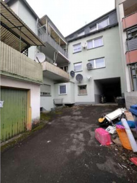 Hofbereich zwischen Wohngebäuden mit grüner Garagentür links und dunklem Garagenzugang rechts, gepflastert, mit Satellitenschüsseln und Müllcontainern