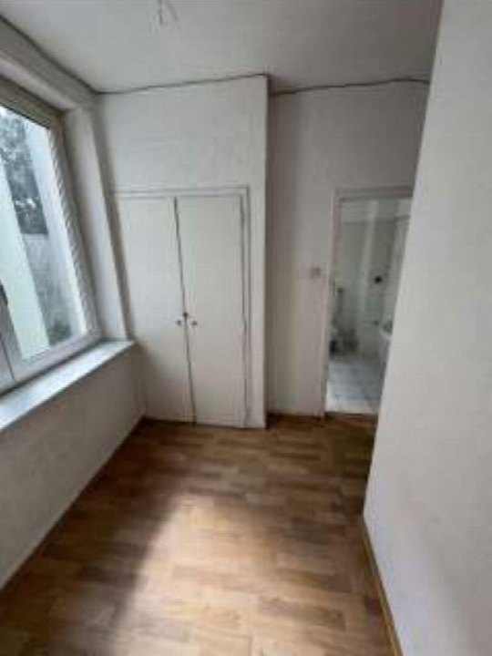 Leerer Raum mit Holzboden, weißer Wand, Fenster und Schrank. Tür führt in angrenzenden Raum