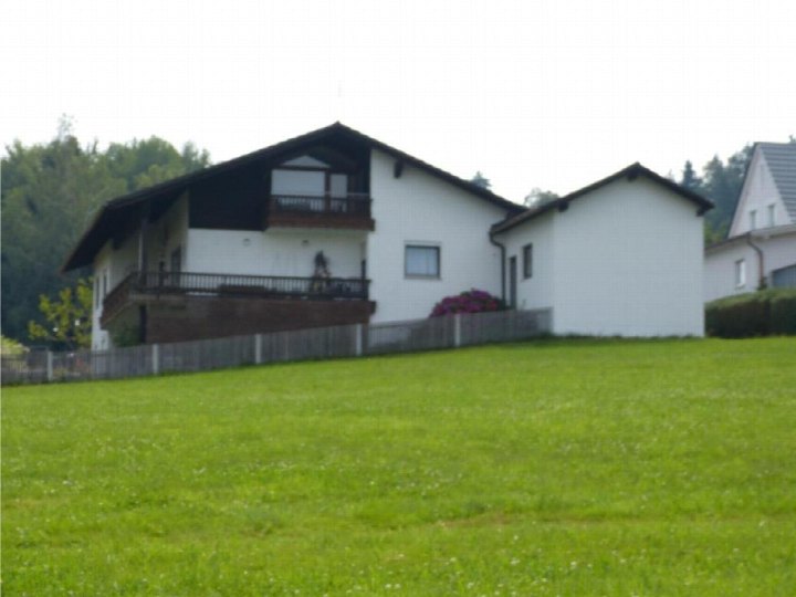 Einfamilienhaus mit dunklem Satteldach, Balkonen und weißer Fassade, umgeben von Grünfläche und Bäumen