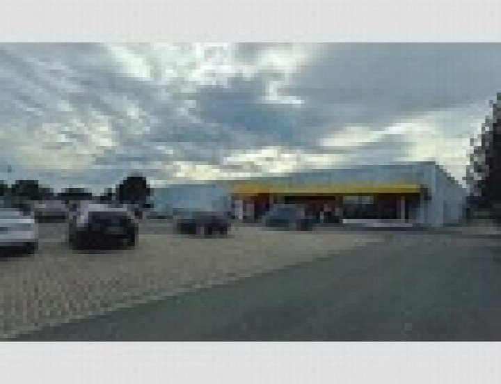 Außenansicht eines Gebäudes mit gelber Markise, geparkten Fahrzeugen und Bäumen im Hintergrund unter bewölktem Himmel