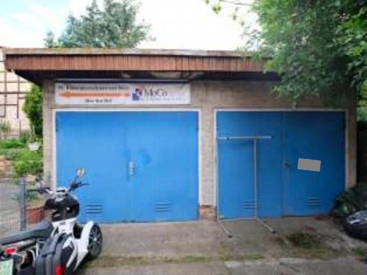 Zwei blaue Garagentore unter einem Holzdach, davor geparkter Roller, im Außenbereich eines Wohn-/Geschäftshauses in Blankenburg