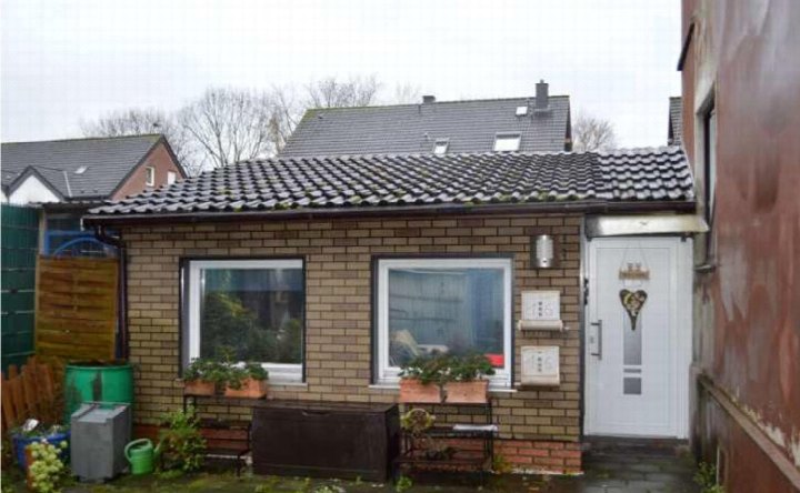Eingangsbereich eines Backsteinhauses mit weißer Tür, Fenstern und Pflanzenkästen. Im Hintergrund weitere Gebäude und ein Zaun