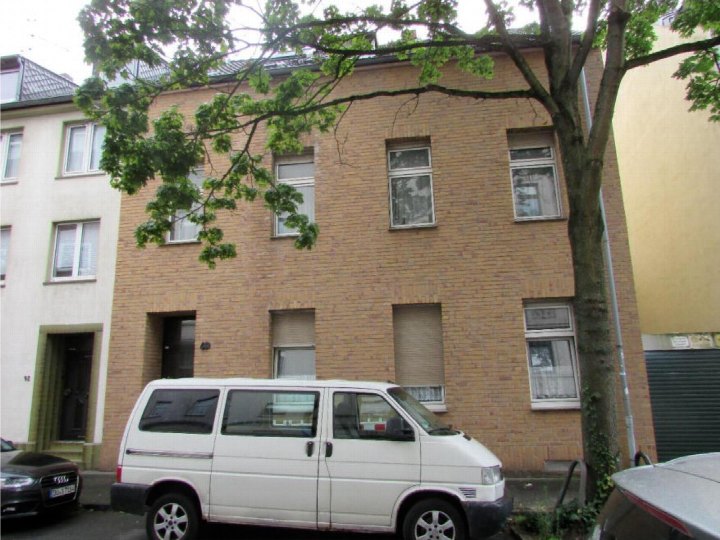 Vorderansicht eines Mehrfamilienhauses aus gelbem Backstein mit Fenstern und Eingang, geparktem weißen Van und Baum im Vordergrund