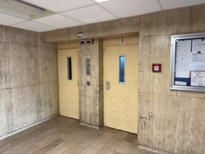 Innenraum mit zwei gelben Aufzugstüren, Betonwänden und Fliesenboden, rechts eine Mitteilungstafel