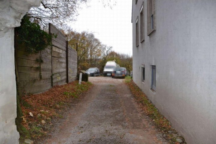 Zugangsweg zu einem Parkplatz mit geparkten Fahrzeugen, flankiert von einer Holzumzäunung und einer Gebäudefassade