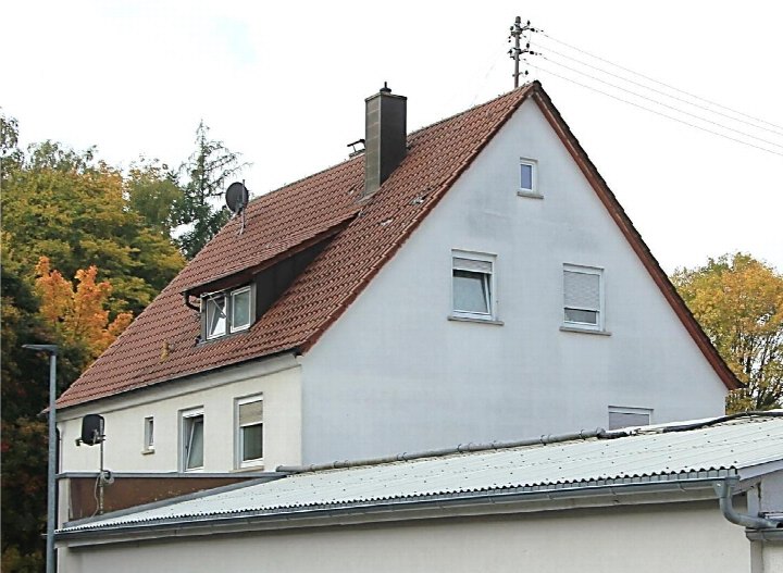 Seitenansicht eines Mehrfamilienhauses mit Satteldach, Dachgauben, Schornstein und Fenstern; im Hintergrund Bäume mit Herbstlaub