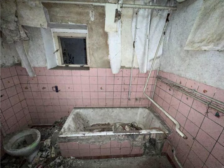 Verfallener Badezimmerbereich mit rosa Fliesen, zerstörter Badewanne und Toilette, sichtbaren Rohrleitungen und Fenster