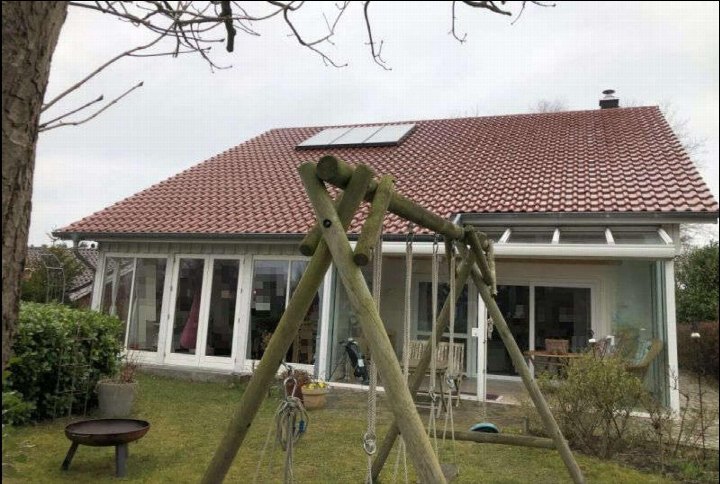 Gartenbereich mit Holzschaukel, Grasfläche, Sträuchern und Feuerschale vor einem Einfamilienhaus mit großflächigen Fenstern und Satteldach