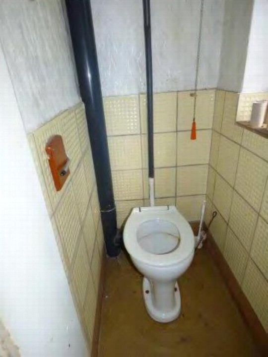 Badezimmer mit weißer Toilette, gelblichem Fliesenbelag und sichtbaren Rohrleitungen an der Wand