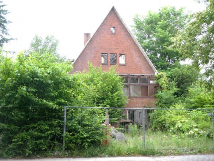 Vorderansicht eines zweistöckigen Backsteinhauses mit Satteldach, umgeben von üppiger Vegetation und einem Metallzaun