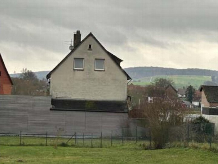 Seitenansicht eines Wohn-/Geschäftshauses mit Satteldach, zwei Oberlichtern und Schornstein, umgeben von Grünfläche und Blick auf Hügel im Hintergrund