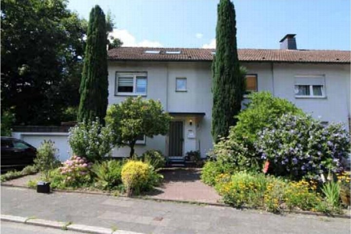 Vorderansicht eines Zweifamilienhauses mit Eingang, Fenstern, Ziegeldach und vorgelagertem Garten mit Sträuchern und Bäumen