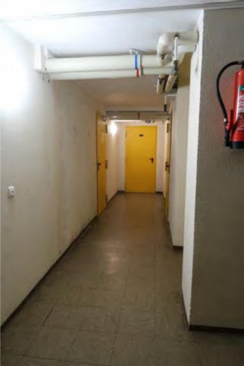 Gewölbter Flur mit Fliesenboden, weißen Wänden, sichtbaren Rohrleitungen und gelber Tür am Ende im Gebäude Alfred-Neumann-Anger 10, München